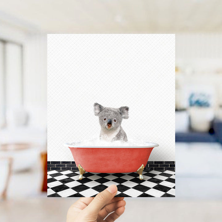 A hand holds up a framed image of a koala bear sitting in a red bathtub filled with bubbles, against a black and white checkered floor.