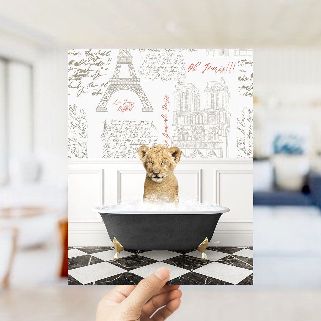 A hand is holding up a printed image featuring a young lion cub sitting in a bathtub filled with water, surrounded by various Paris-themed illustrations.