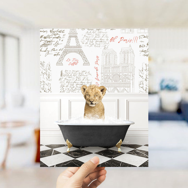A hand is holding up a printed image featuring a young lion cub sitting in a bathtub filled with water, surrounded by various Paris-themed illustrations.