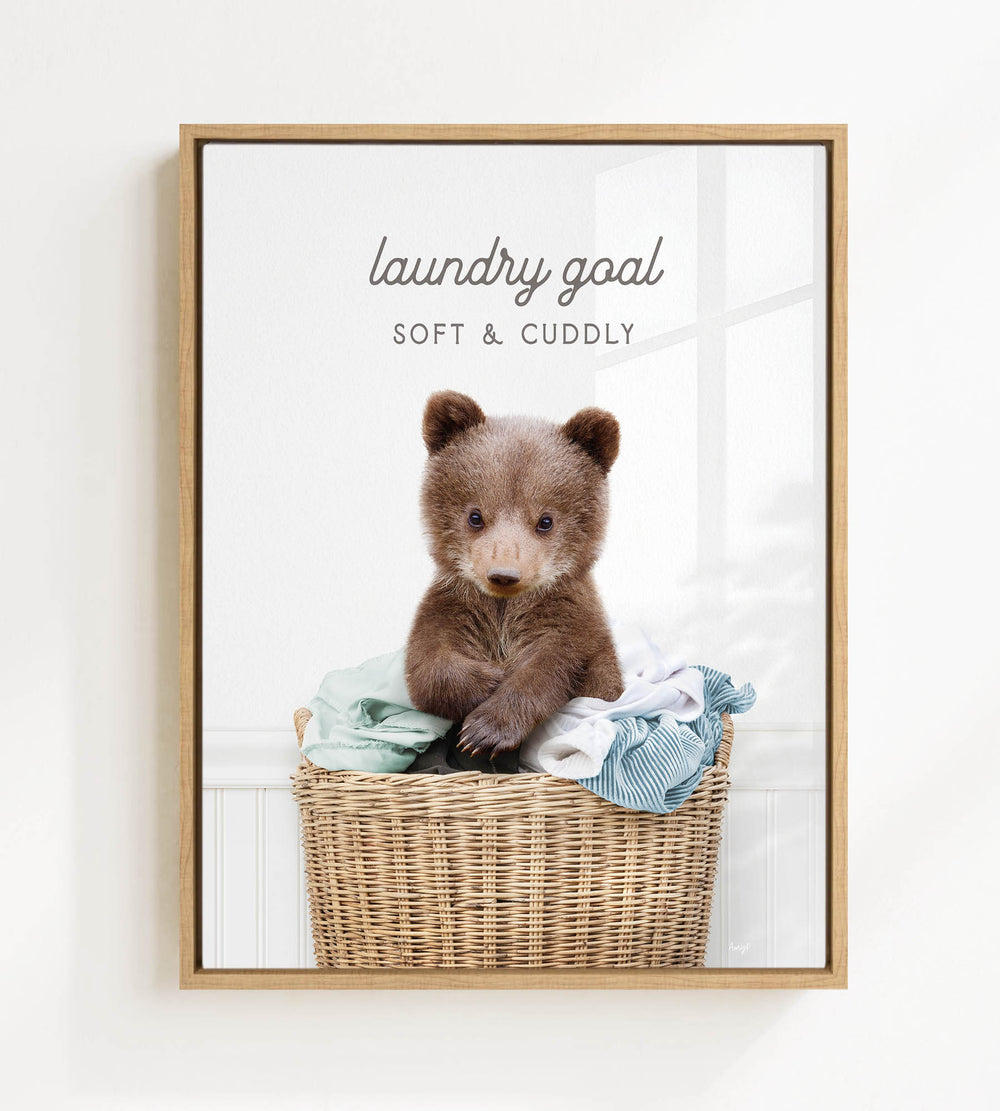 A cute brown bear sitting in a wicker laundry basket filled with clothes, with the text "laundry goal" and "soft & cuddly" written above it.