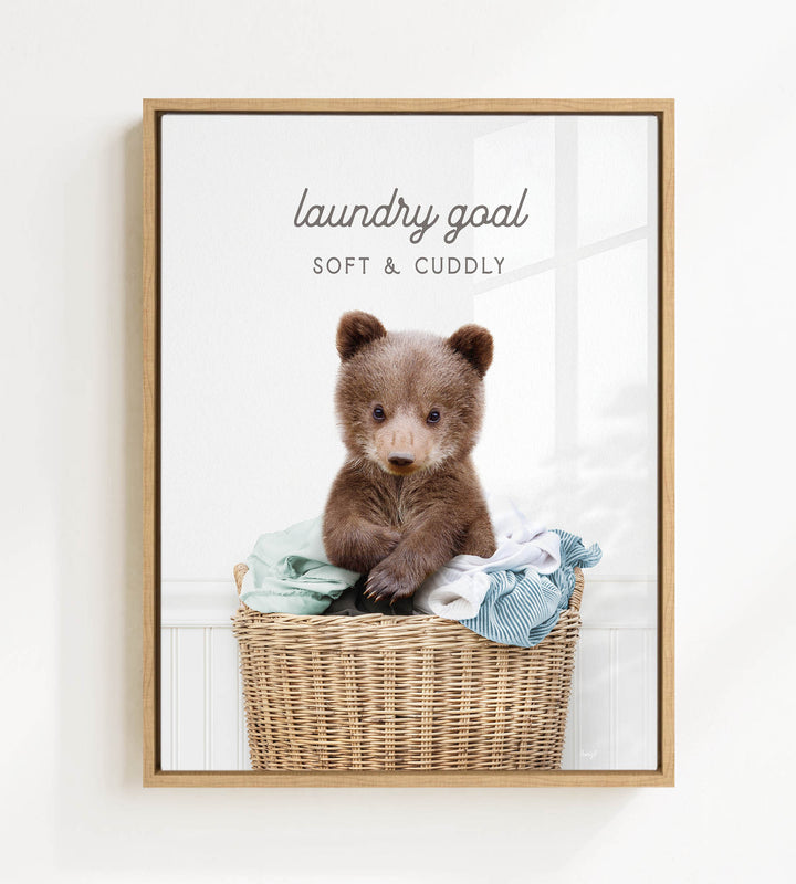 A cute brown bear sitting in a wicker laundry basket filled with clothes, with the text "laundry goal" and "soft & cuddly" written above it.