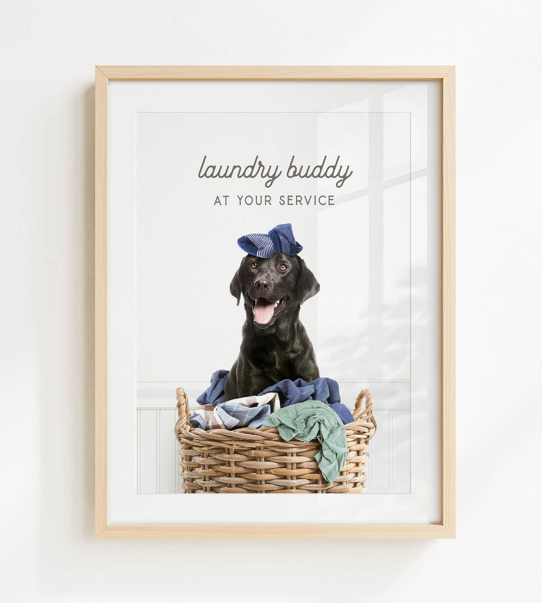 A black dog sitting in a wicker laundry basket, wearing a blue hat and surrounded by clothes.