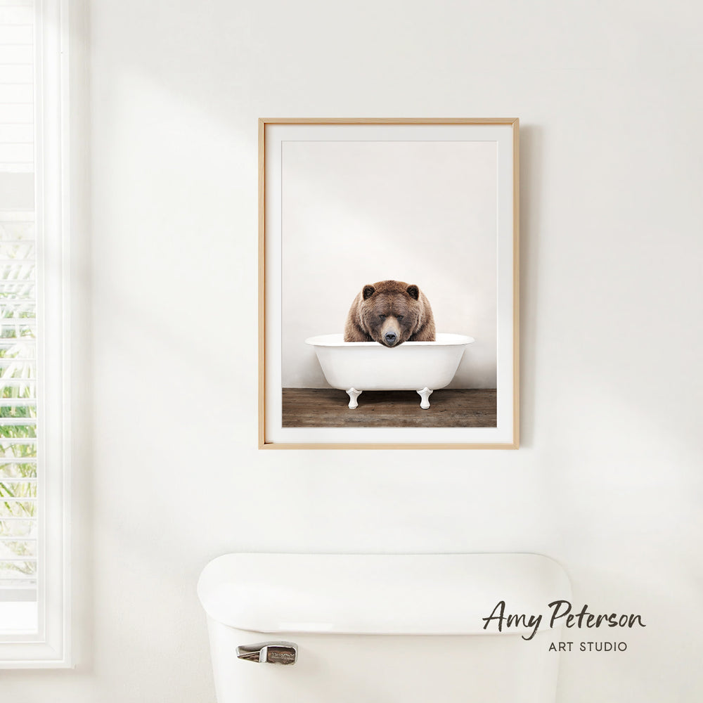 A framed image of a bear sitting in a white bathtub is hanging on a white wall in a bathroom.