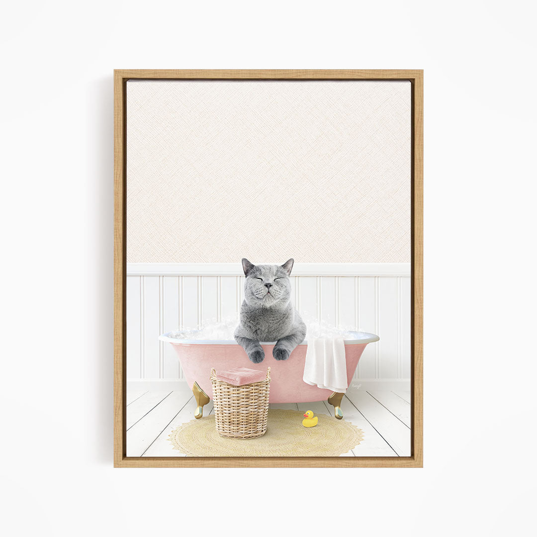 A gray cat sitting in a pink bathtub filled with bubbles, with a basket of bath toys nearby.