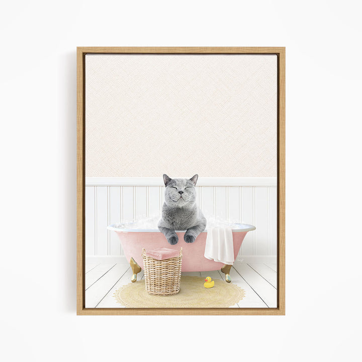A gray cat sitting in a pink bathtub filled with bubbles, with a basket of bath toys nearby.