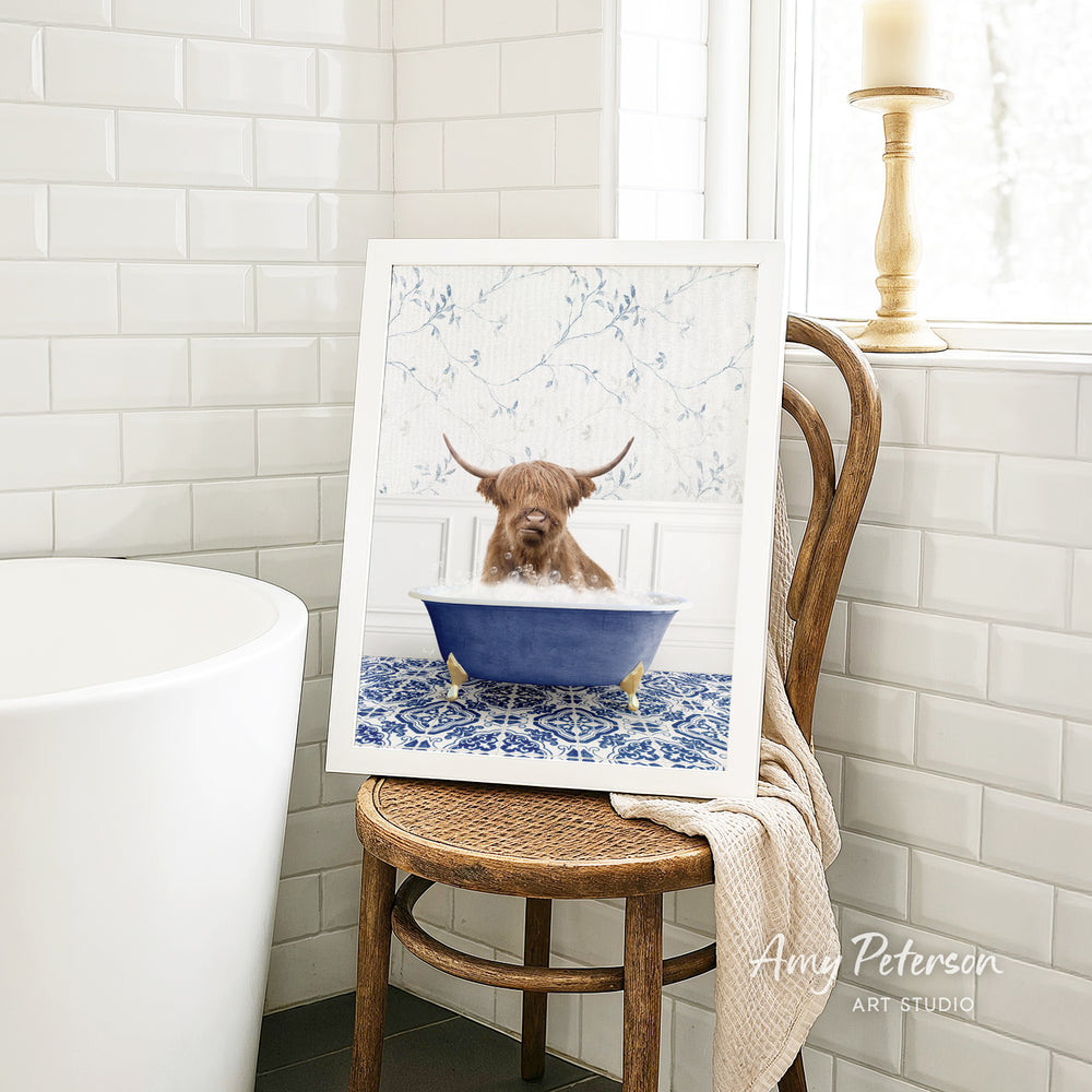 A framed image of a Highland cow sitting in a blue bathtub, surrounded by a white and blue floral pattern, is displayed on a wooden chair in a bathroom setting.