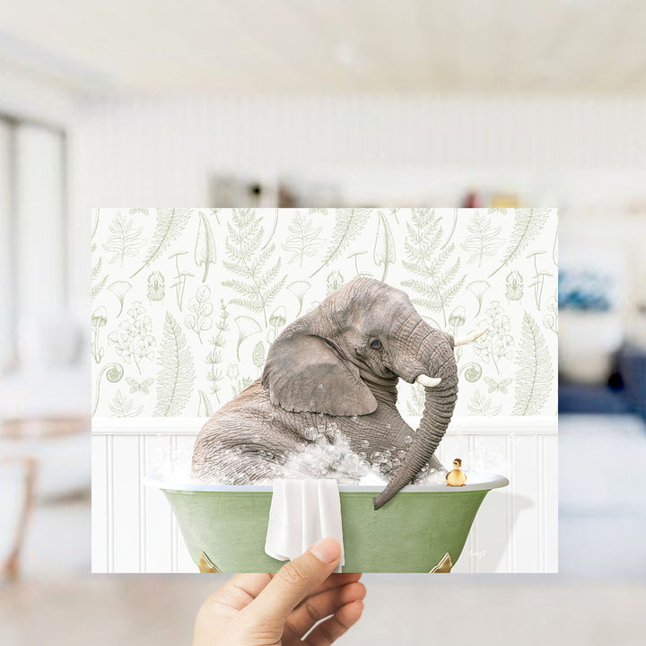 A hand holds up a framed image depicting an elephant sitting in a green bathtub, surrounded by a fern-patterned wall.
