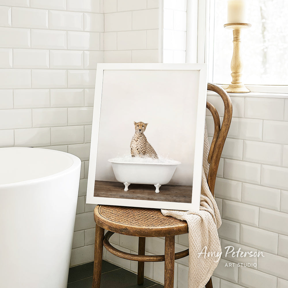A framed image of a cheetah sitting in a bathtub is displayed on a wooden chair in a bathroom setting.