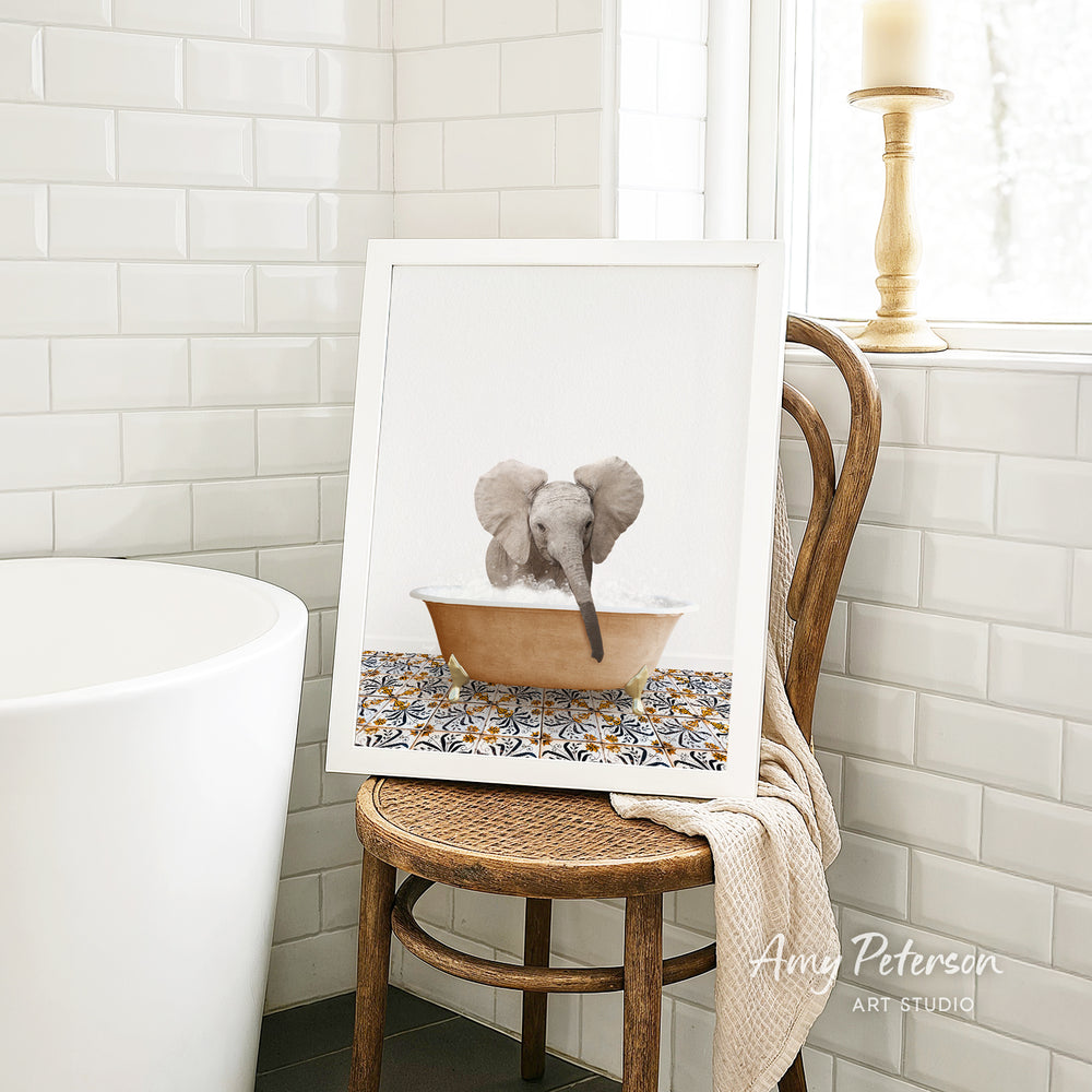 A framed image of a baby elephant sitting in a bathtub, surrounded by a patterned tile design, is displayed on a wooden chair in a bathroom setting.