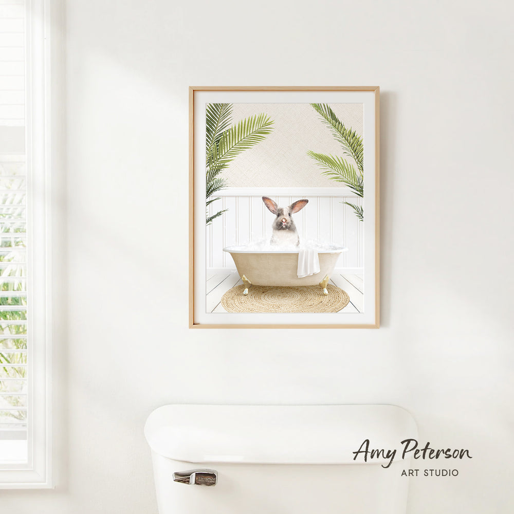 A framed print of a rabbit sitting in a bathtub, surrounded by palm leaves, hangs on a white wall in a bathroom.
