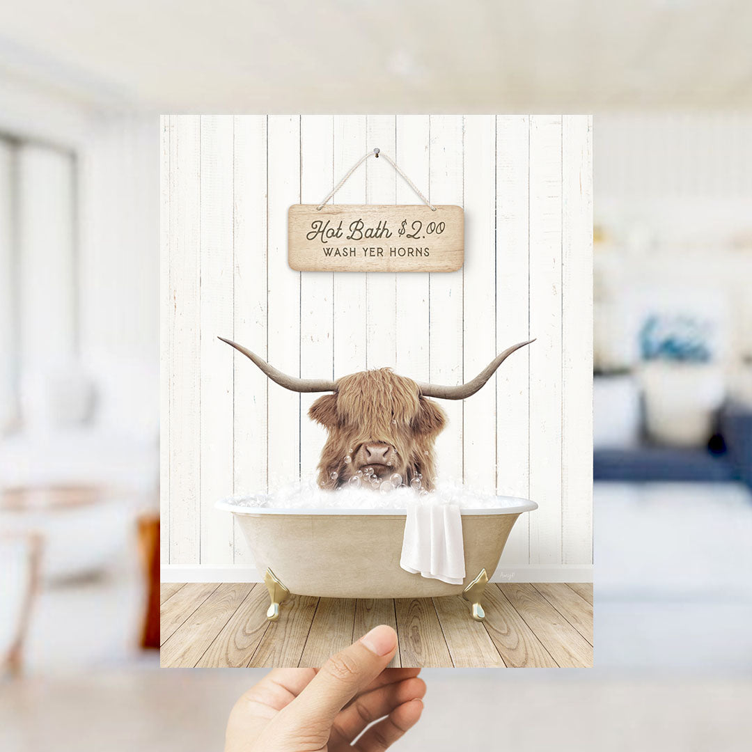 A hand holding a framed image of a horned animal, likely a yak or a similar animal, sitting in a bathtub filled with water. The sign above the bathtub reads "Hot Bath $2.00 WASH YOUR HORSES".