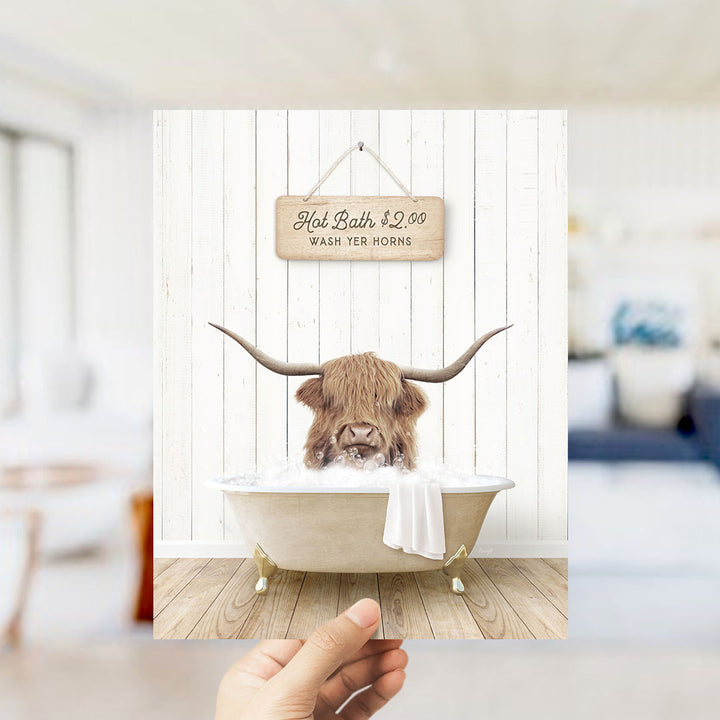 A hand holding a framed image of a horned animal, likely a yak or a similar animal, sitting in a bathtub filled with water. The sign above the bathtub reads "Hot Bath $2.00 WASH YOUR HORSES".