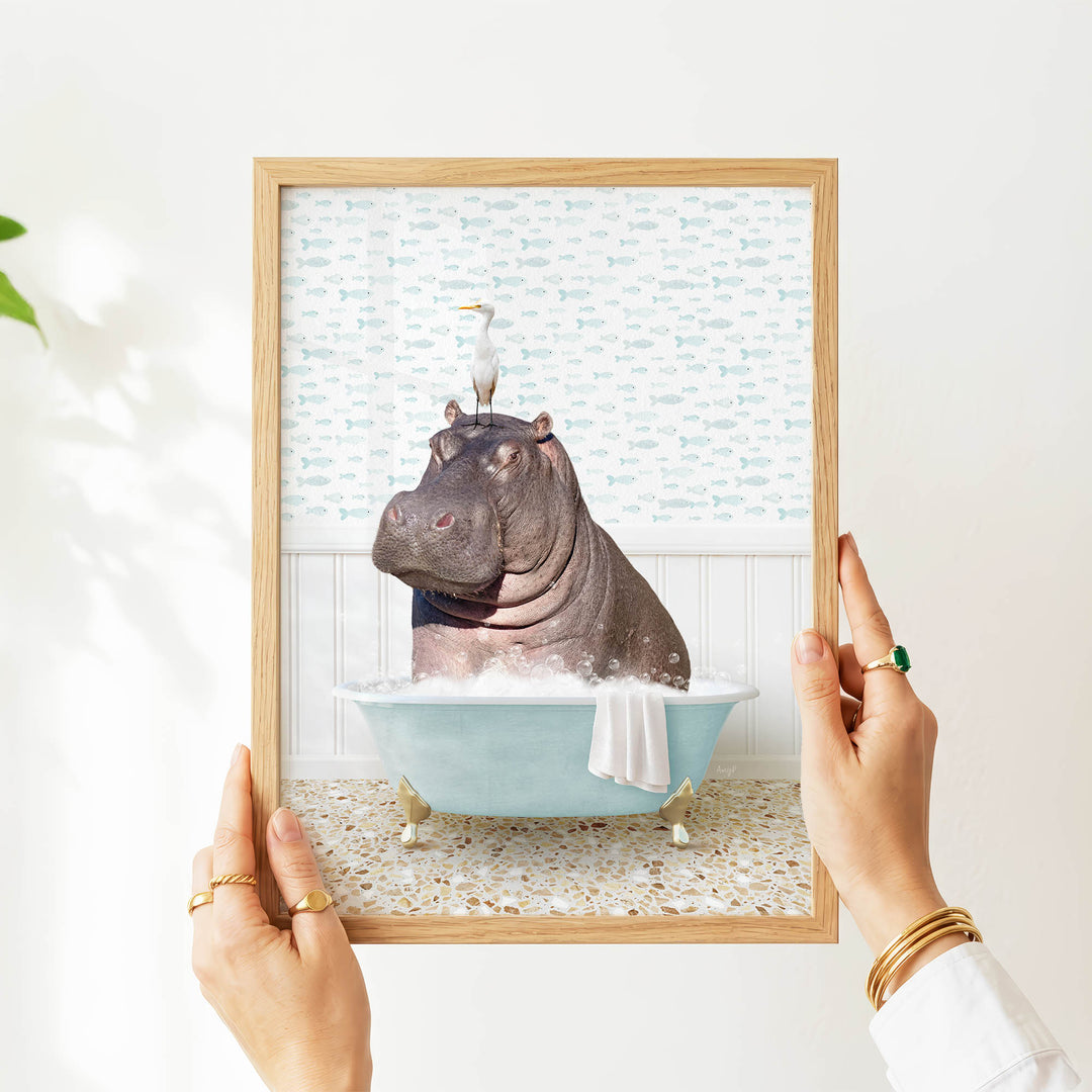 A person's hands are holding a framed image of a hippopotamus sitting in a bathtub filled with water and bubbles.