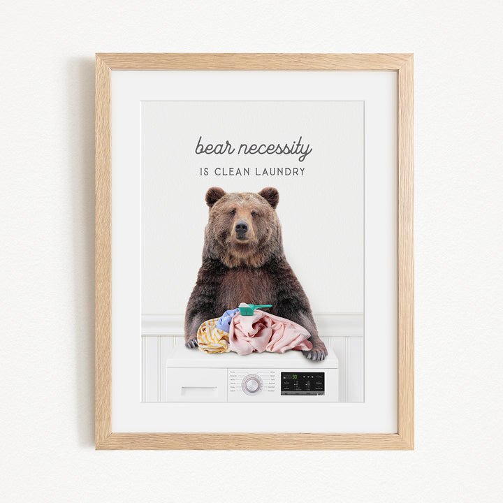 A framed image of a brown bear sitting on a washing machine, with the text "bear necessity is clean laundry" above it.