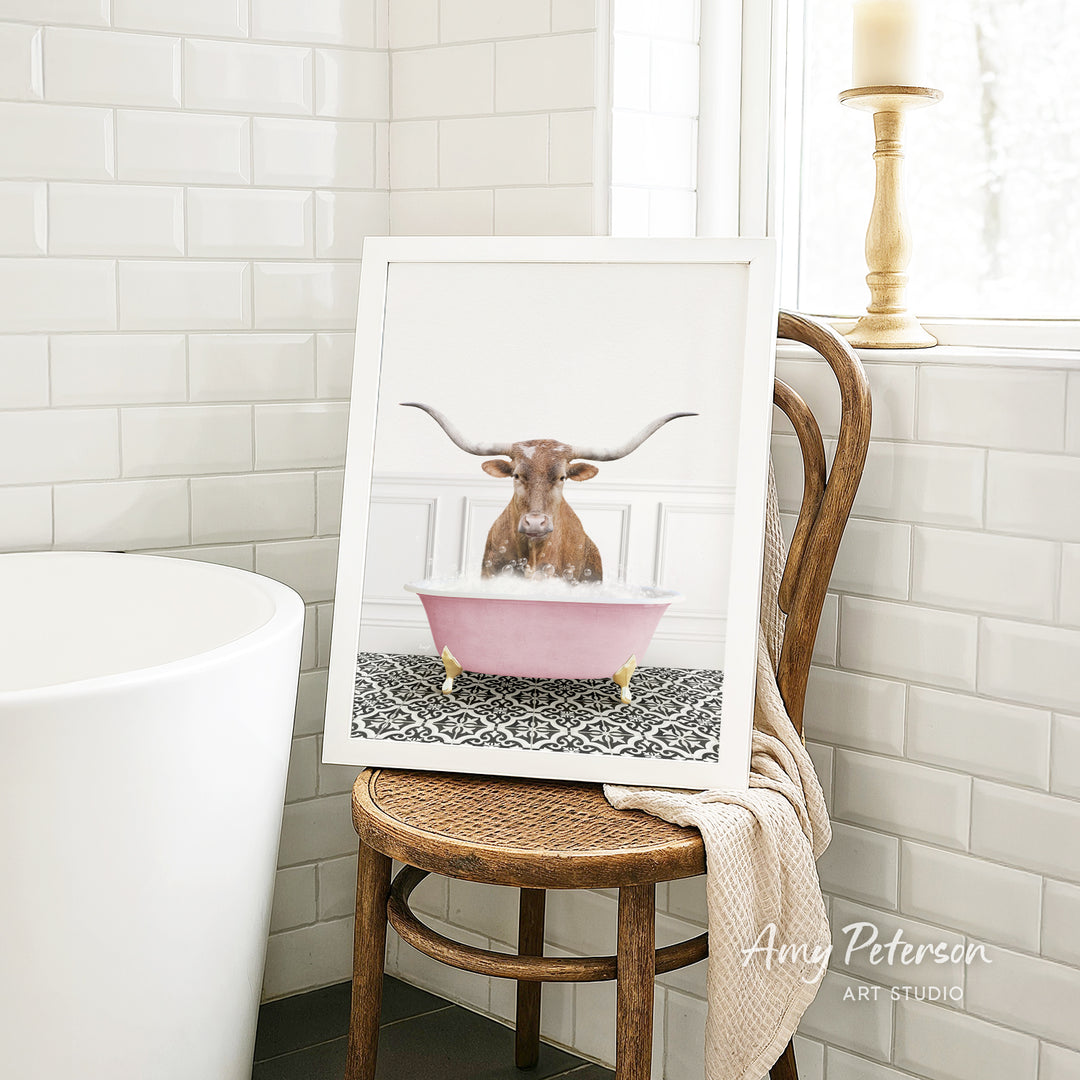 A framed image of a bull with horns sitting in a pink bathtub, surrounded by a patterned rug, is displayed on a wooden chair in a bathroom setting.
