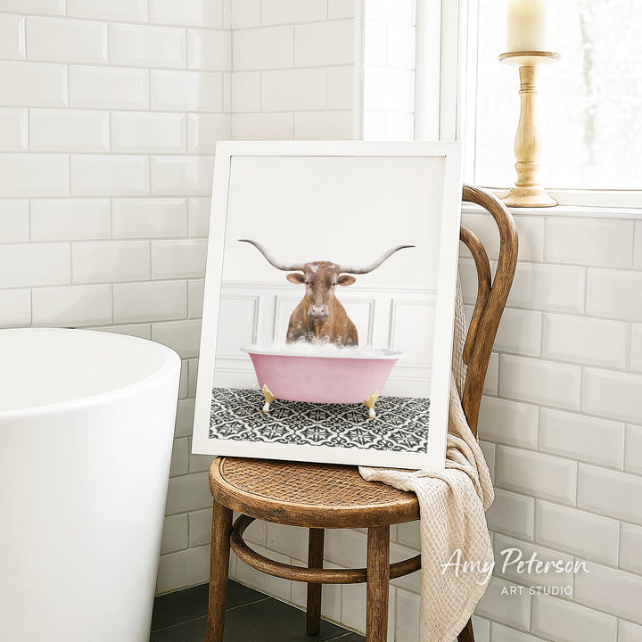 A framed image of a bull with horns sitting in a pink bathtub, surrounded by a patterned rug, is displayed on a wooden chair in a bathroom setting.