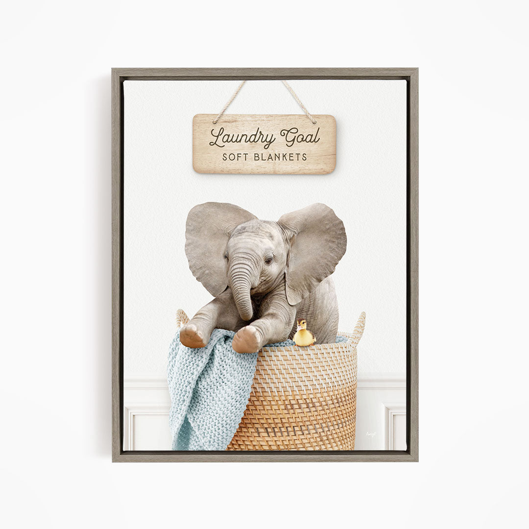 A framed image of an elephant sitting in a woven basket, with a sign above it that says "Laundry Goal Soft Blankets".