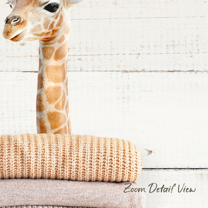 A giraffe standing behind a stack of beige and brown textured blankets, with a white wooden background.