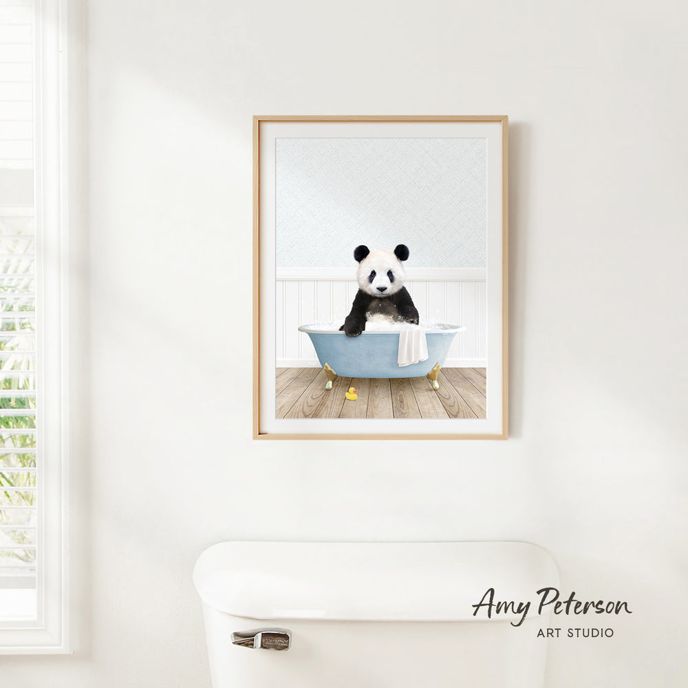 A framed image of a panda sitting in a bathtub, with a towel draped over its body, is hanging on a white wall.