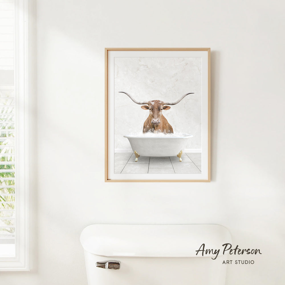 A framed image of a bull with long horns sitting in a bathtub, with a white toilet in the foreground.