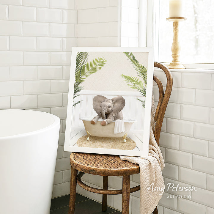 A framed image of two elephants sitting in a bathtub, surrounded by green leaves, is displayed on a wooden chair in a bathroom setting.
