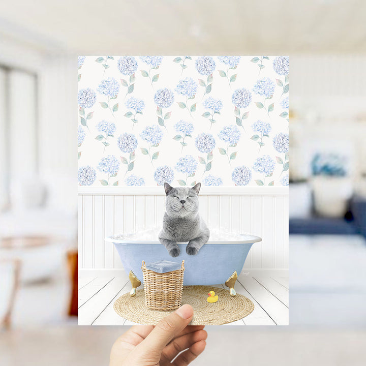 A gray cat sitting in a blue bathtub filled with bubbles, surrounded by a wicker basket and a rubber duck.