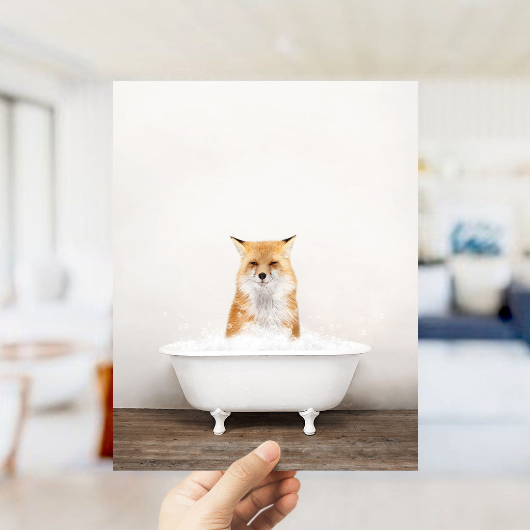 A hand holds up a framed image of a fox sitting in a white bathtub filled with bubbles.