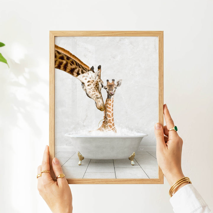 A person's hands are holding a framed picture of two giraffes in a bathtub, with one giraffe appearing to be kissing the other giraffe.