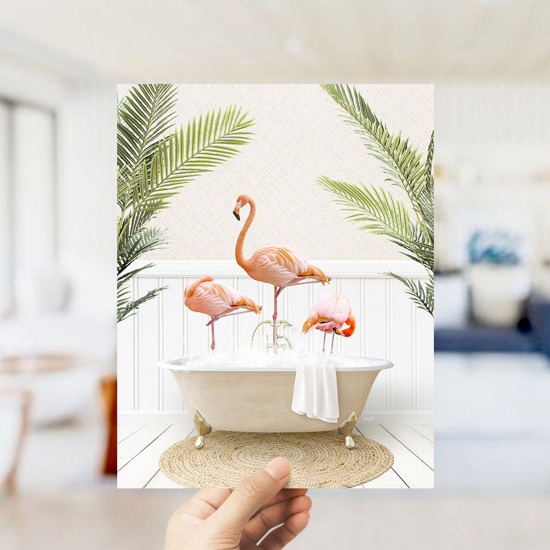 A hand holds up a framed image depicting three flamingos standing in a bathtub filled with water, surrounded by palm leaves.