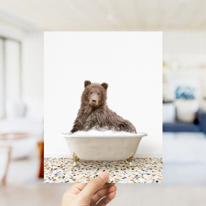 A brown bear sitting in a white bathtub filled with bubbles, with a hand holding the photo up to the camera.