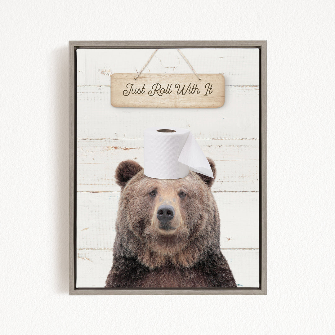 A framed image of a brown bear wearing a roll of toilet paper on its head, with a wooden sign above it that says "Just Roll With It".