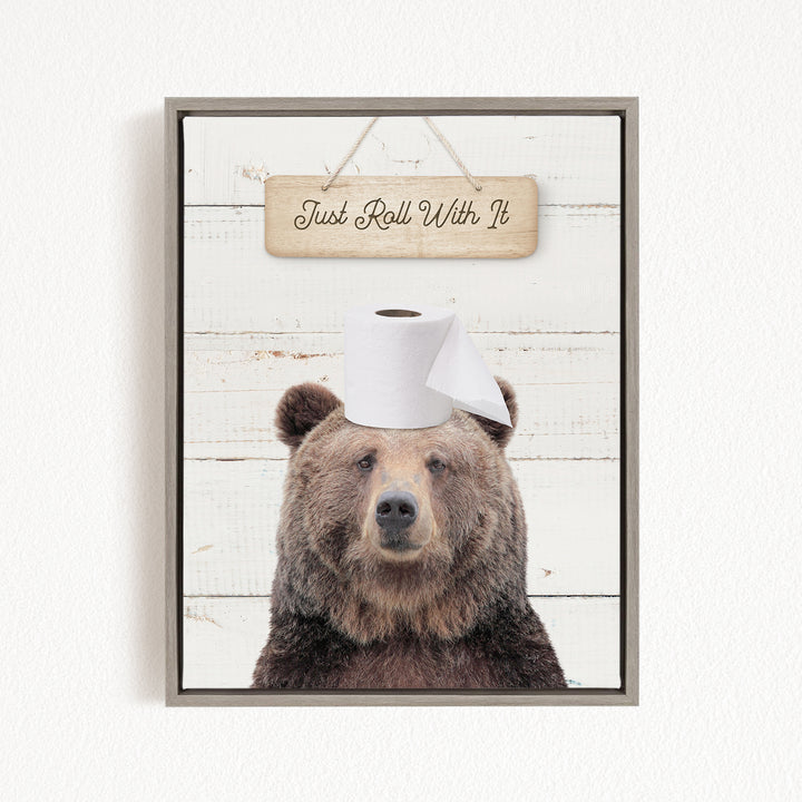 A framed image of a brown bear wearing a roll of toilet paper on its head, with a wooden sign above it that says "Just Roll With It".