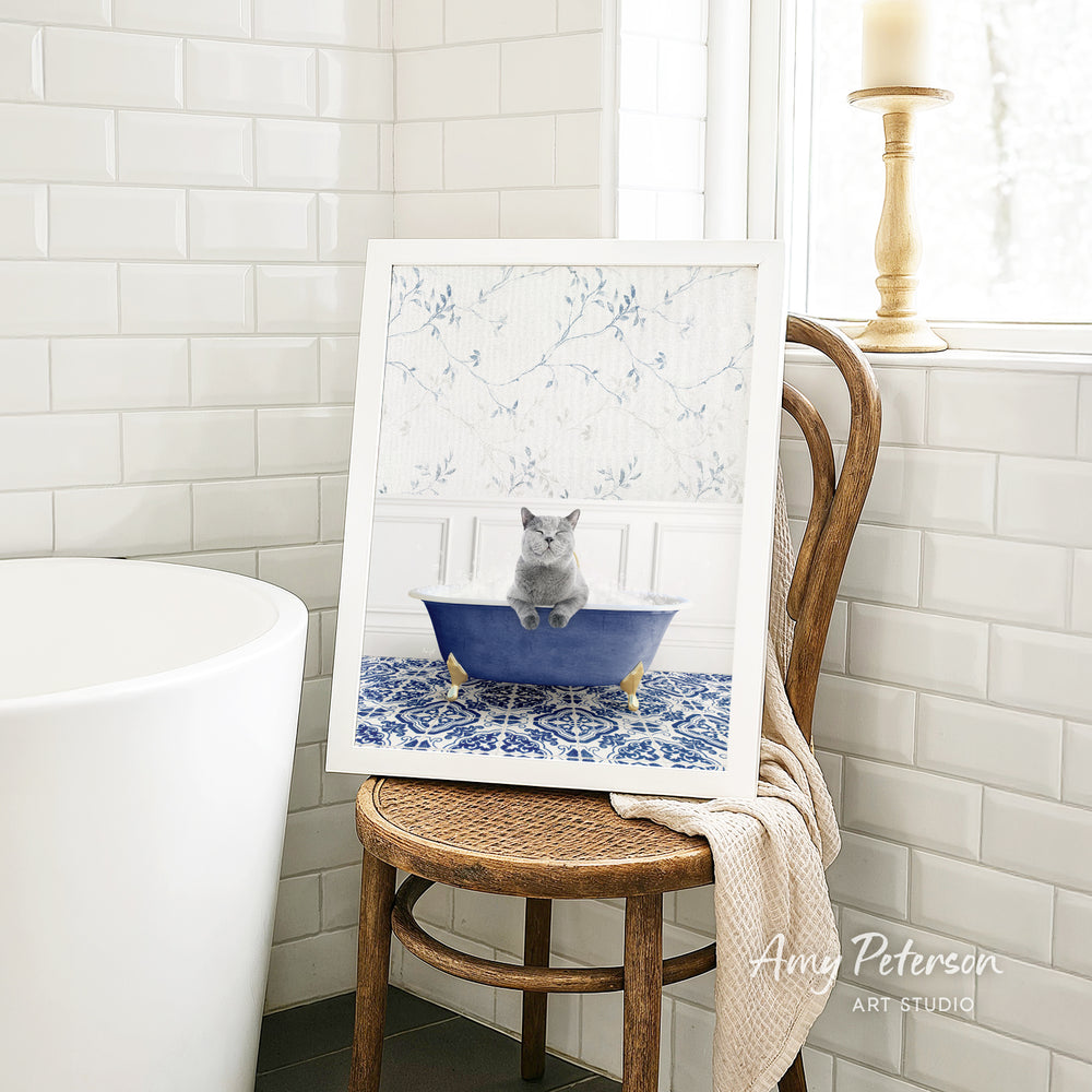 A framed image of a gray cat sitting in a blue bathtub, with a white floral pattern on the wall behind it, is displayed on a wooden chair in a bathroom setting.