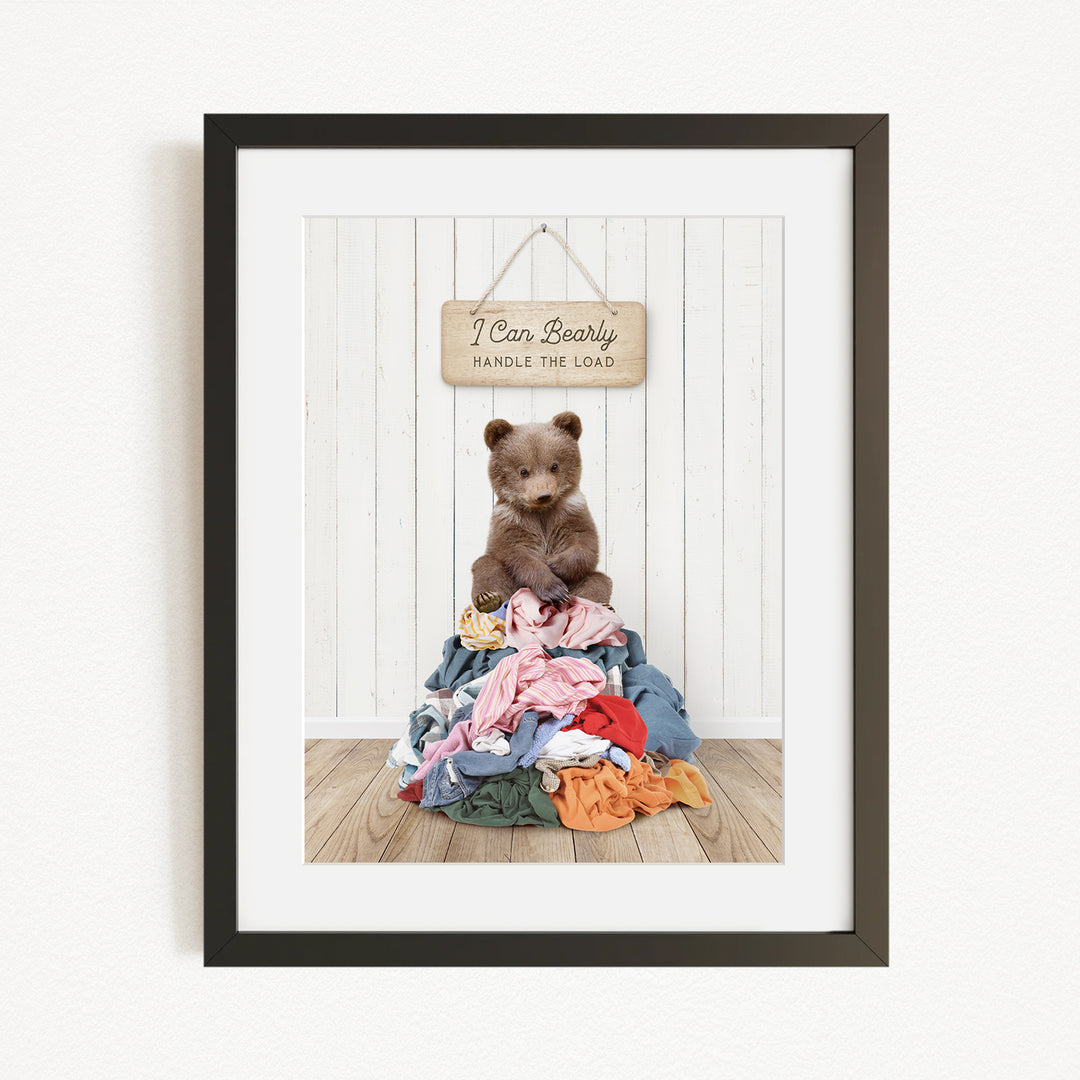 A framed image of a teddy bear sitting atop a pile of clothes, with a wooden sign above that reads "I Can Bearly Handle the Load".