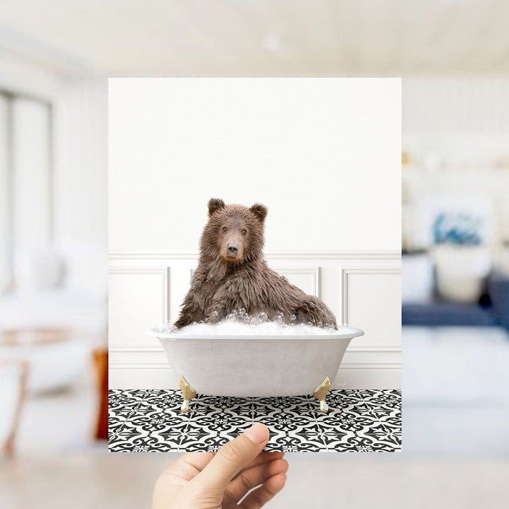 A brown bear sitting in a white bathtub filled with bubbles, with a hand holding the image up to the viewer.