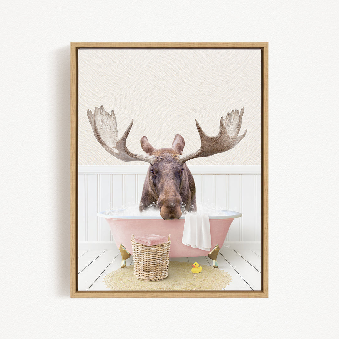A moose with large antlers is sitting in a pink bathtub filled with water, surrounded by a basket of rubber ducks and a towel.