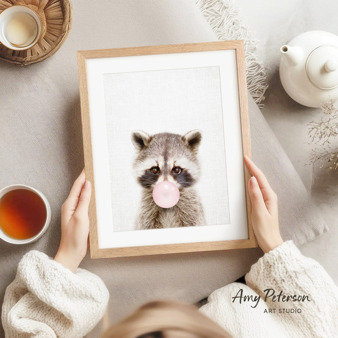 A person is holding a framed picture of a raccoon blowing a bubble with a pink bubble gum bubble, with a teapot, a cup of tea, and a bowl of tea nearby.