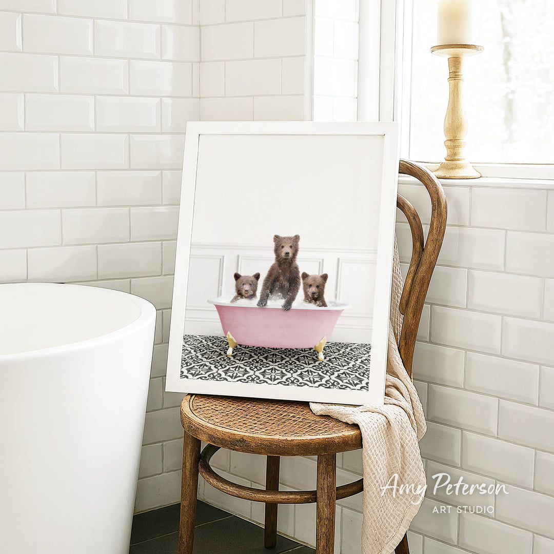 A framed image of three bears sitting in a pink bathtub, with a towel draped over the back of a wooden chair in a bathroom setting.