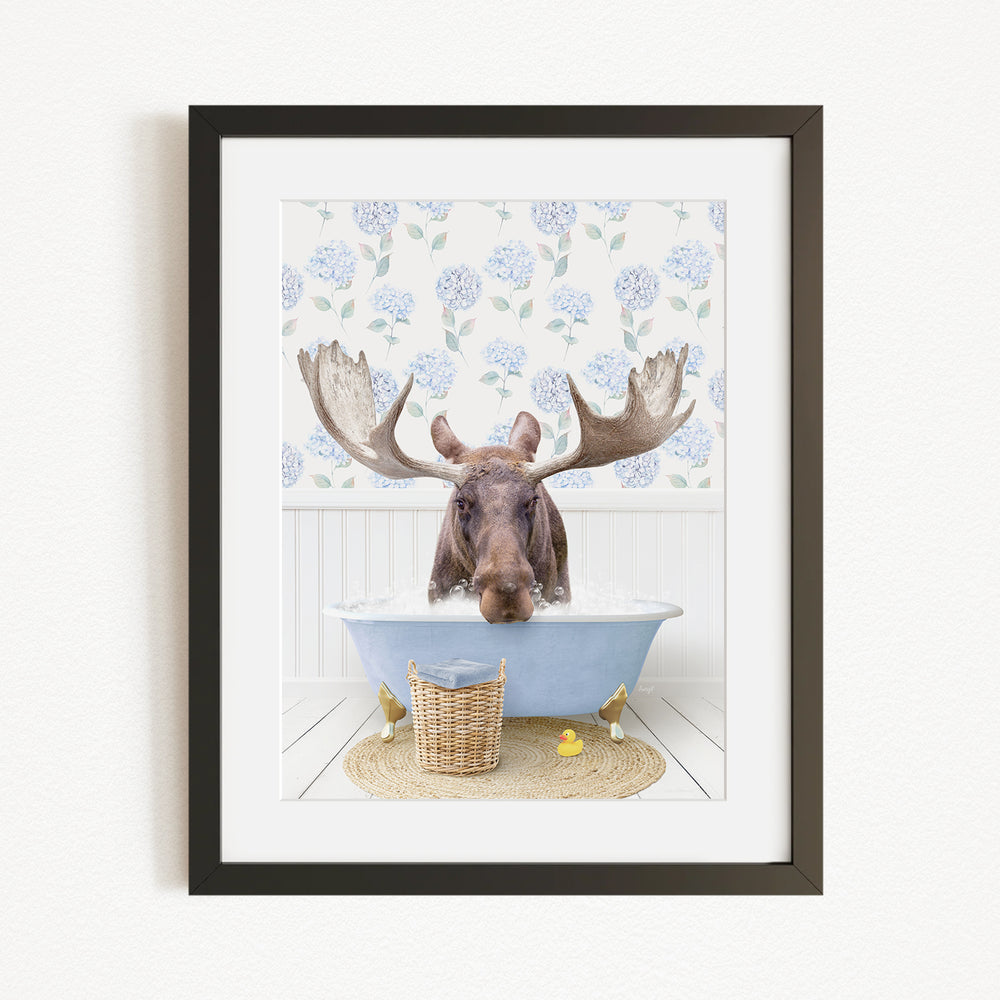 A moose with large antlers is sitting in a blue bathtub, surrounded by a floral patterned wall and a basket of rubber ducks.
