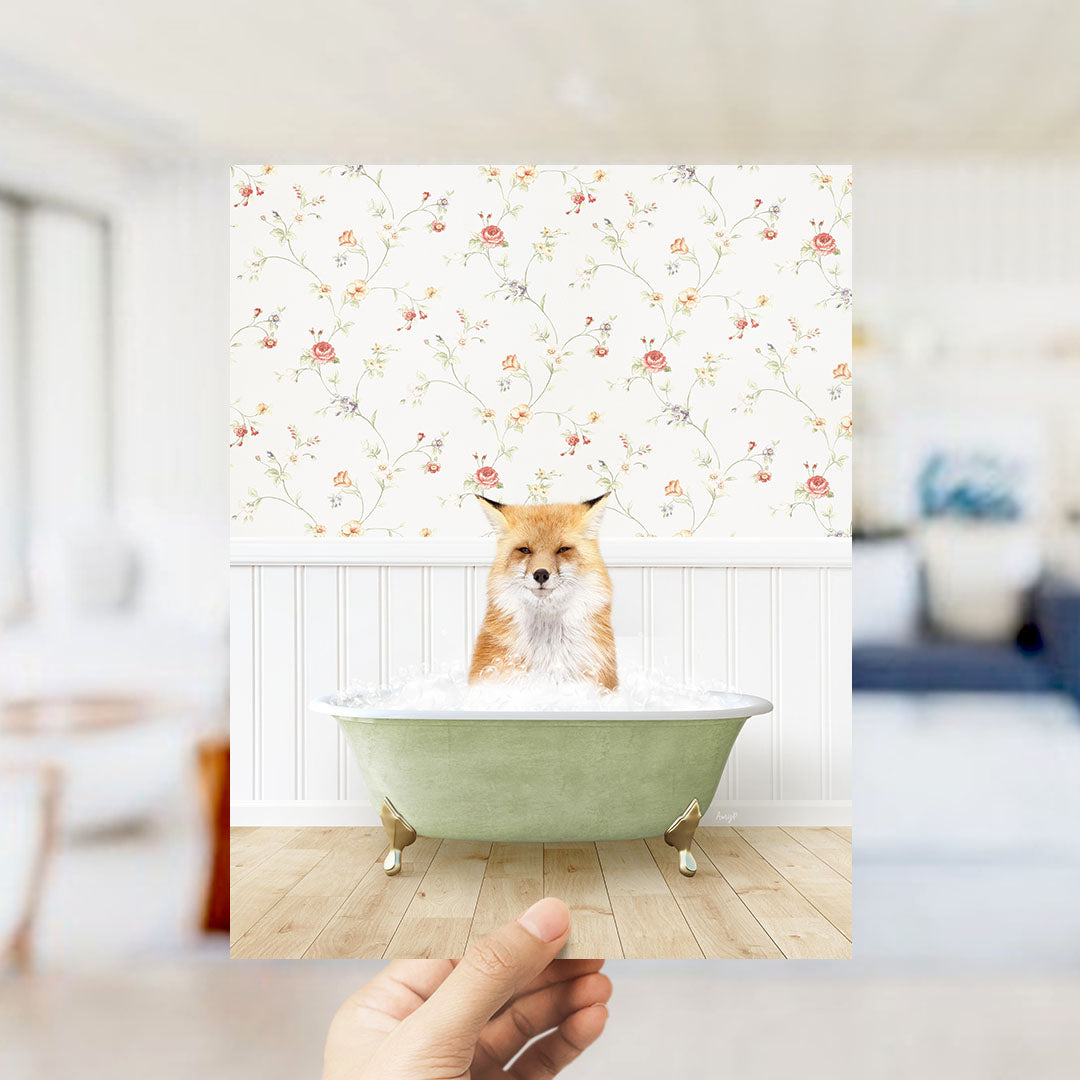 A hand holds up a framed image of a fox sitting in a green bathtub filled with bubbles, with a floral wallpaper in the background.