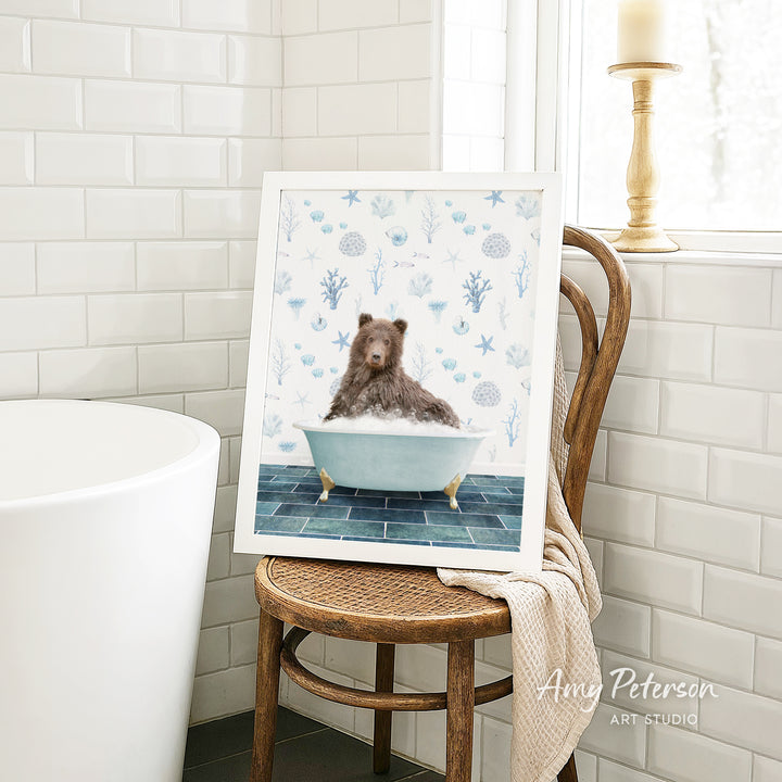 A framed image of a bear sitting in a bathtub, surrounded by blue and white floral patterns, is displayed on a wooden chair in a bathroom setting.