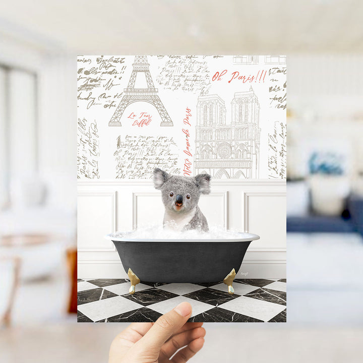 A hand is holding up a framed image of a koala bear sitting in a bathtub filled with water, surrounded by various Paris-themed illustrations and text.