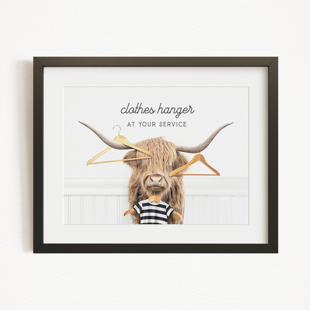 A framed image depicting a large, furry animal with long horns, wearing a striped shirt and holding a pair of clothes hangers, with the text "clothes hanger at your service" above it.