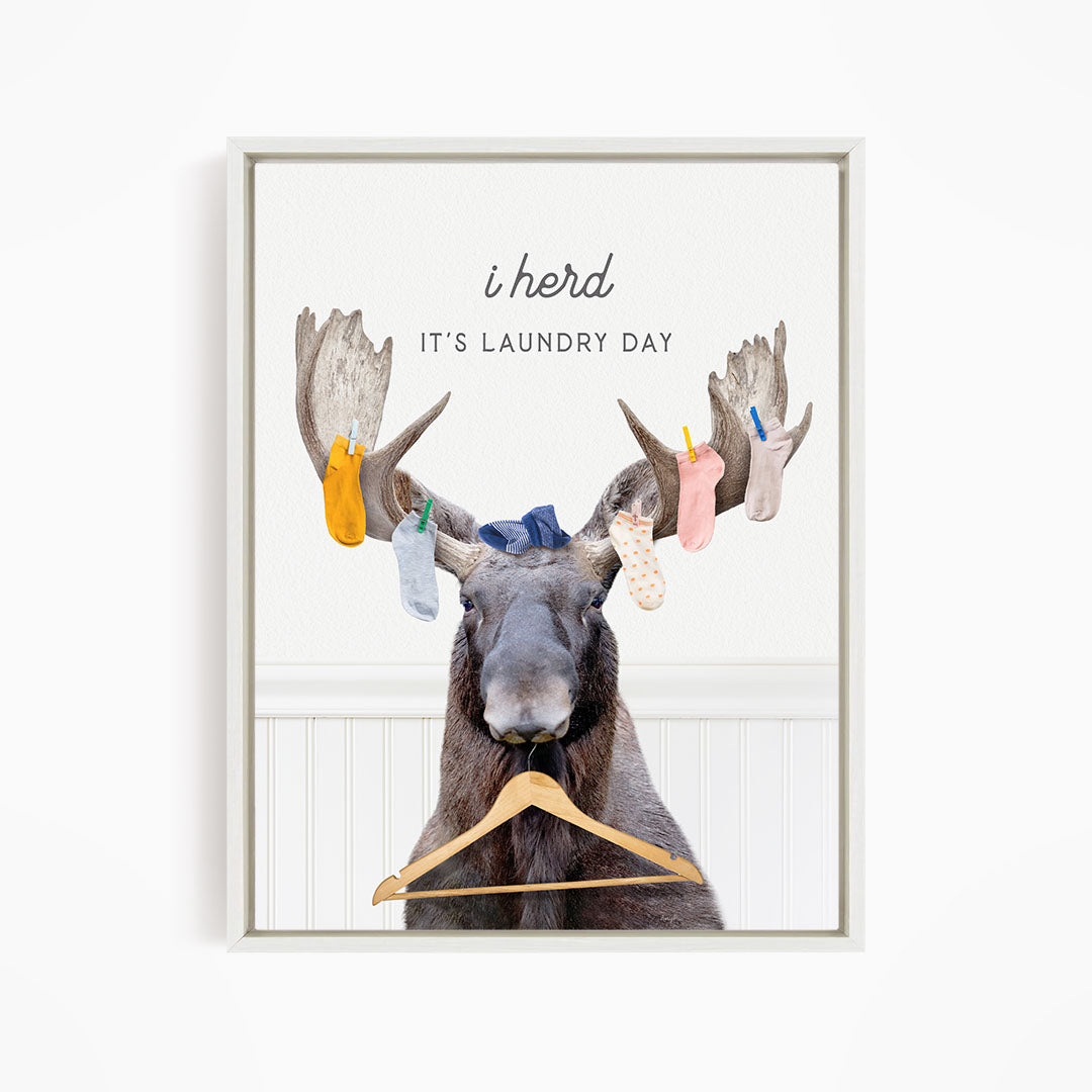 A framed print featuring a cartoon image of a moose with a pile of clothes hanging from its antlers, accompanied by the text "It's laundry day" and the word "herd".