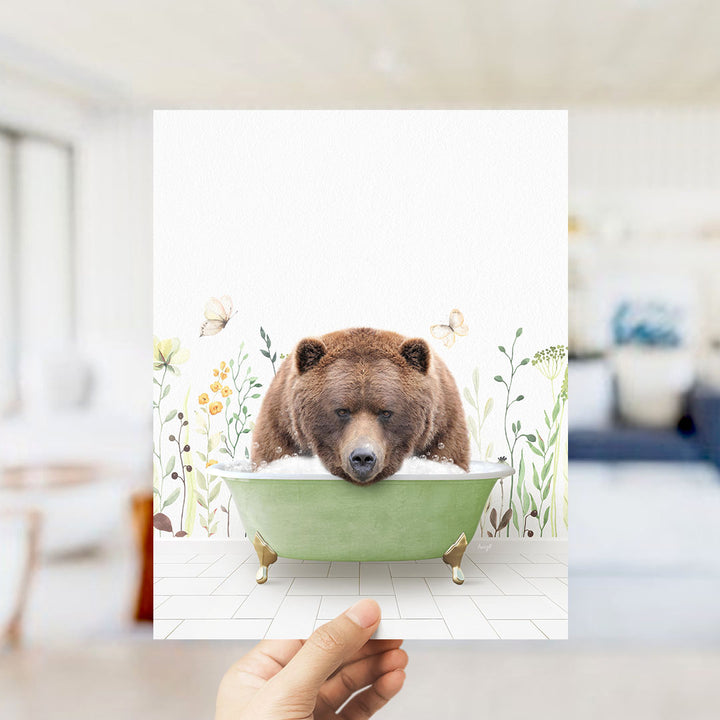 A hand holds up a framed image depicting a brown bear sitting in a green bathtub, surrounded by flowers and butterflies.