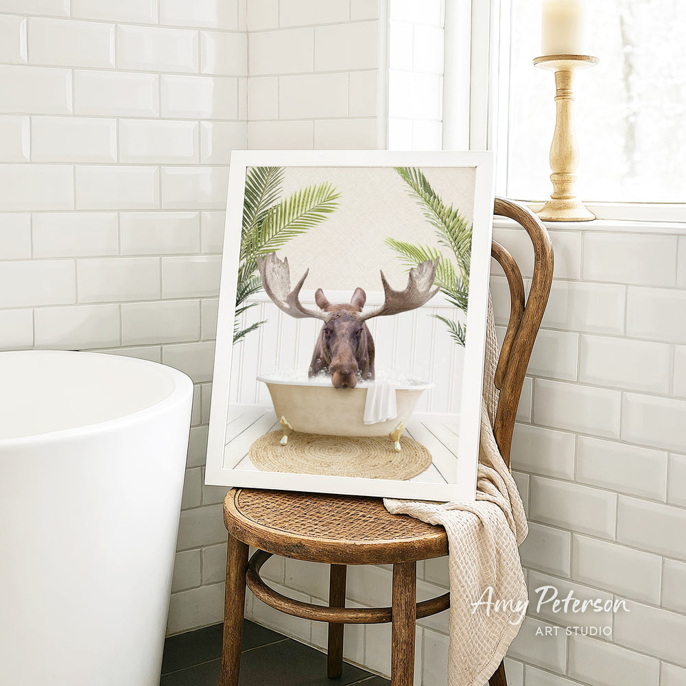 A framed image of a moose sitting in a bathtub, surrounded by tropical plants, is displayed on a wooden chair in a bathroom setting.