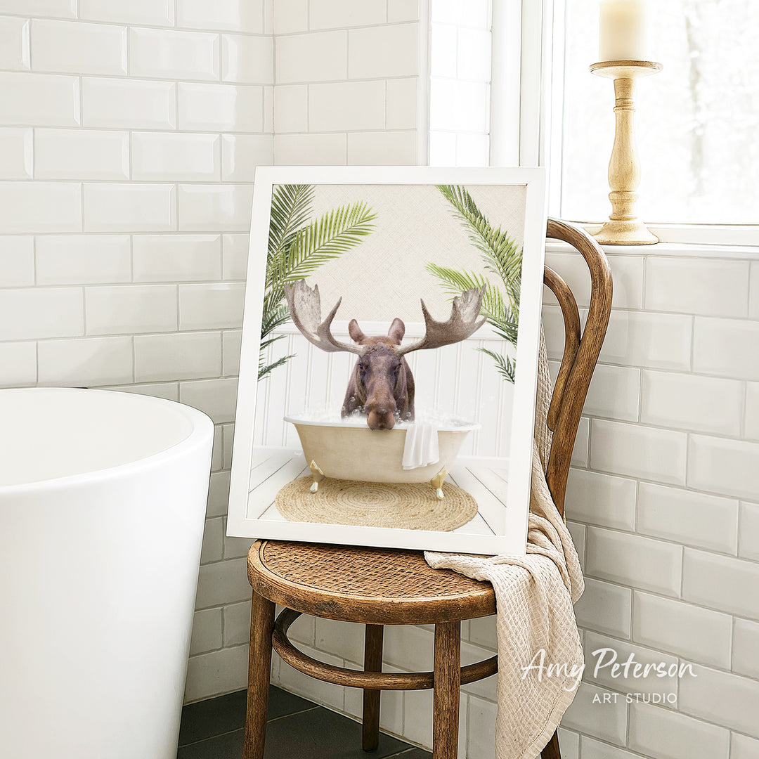 A framed image of a moose sitting in a bathtub, surrounded by tropical plants, is displayed on a wooden chair in a bathroom setting.