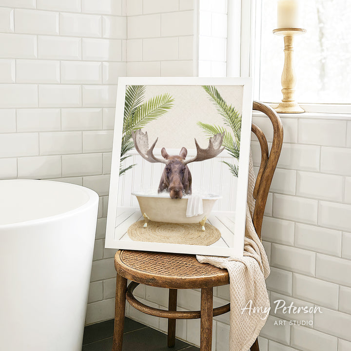 A framed image of a moose sitting in a bathtub, surrounded by tropical plants, is displayed on a wooden chair in a bathroom setting.