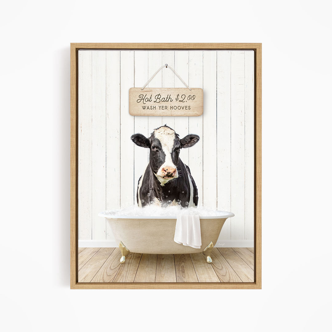 A black and white cow is sitting in a white bathtub, with a wooden sign above it that reads "Hot Bath $2.00 WASH YER BOOOVES".