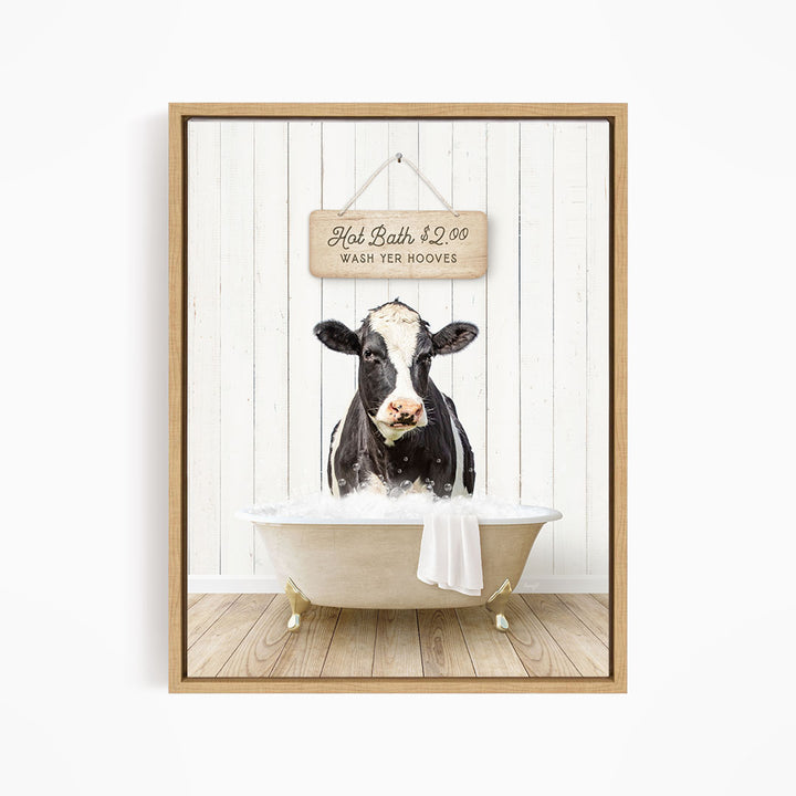 A black and white cow is sitting in a white bathtub, with a wooden sign above it that reads "Hot Bath $2.00 WASH YER BOOOVES".